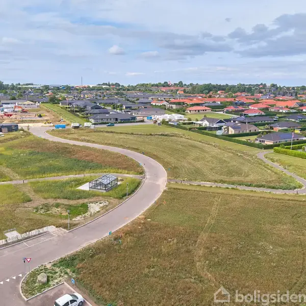 Fremtidig byggeplads vises fra oven, med en snoet vej gennem åbne græsarealer omgivet af nybyggeri og boligområder. Distant horisont med træer og skyet himmel skaber en fredfyldt scene.
