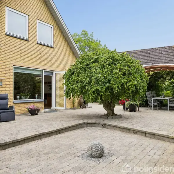Gul murstenshus har en terrasse med fliser og et centralt træ. En afslapningsstol og blomsterpotter står ved husets vindue. I baggrunden er et overdækket siddeområde med havemøbler.