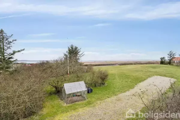 Et lille drivhus står stille i en frodig have omgivet af træer med en stor, åben græsplæne og udsigt til et fladt landskab i baggrunden.