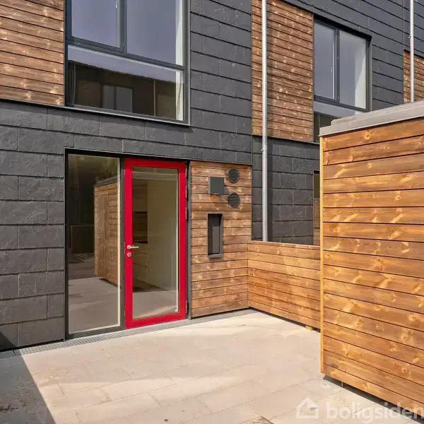 En moderne facade viser en sort bygning med store vinduer og en rød dør. Området har træbeklædning og en flisebelagt gårdhave, hvilket giver et stilrent udtryk.