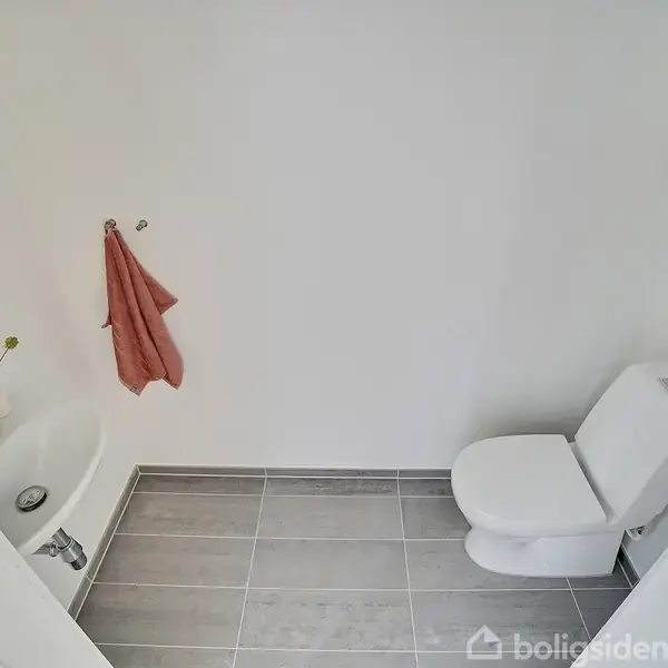 Toilet står ved højre væg uden aktivitet. Flisegulv og hvid væg omgiver rummet. Vask ved venstre væg med vaser og rødt håndklæde hængende ved håndvasken uden nogen synlig tekst.