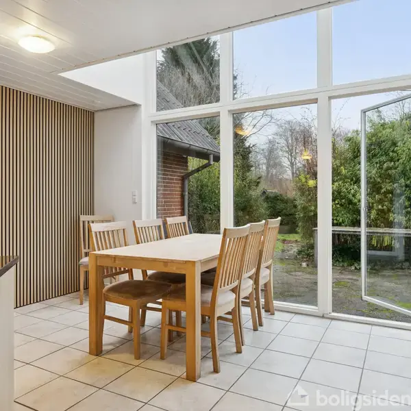 Et spisebord med otte træstole står i et lyst rum med store vinduer, der åbner ud til en have. Flisegulv og lyse træpaneler fuldender indretningen.