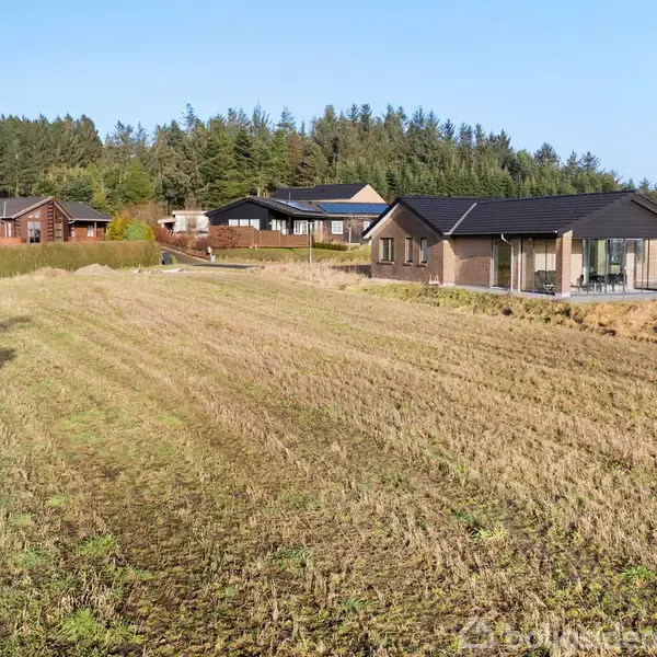 Hus ligger på en stor, græsklædt grund omgivet af åbne marker og flere huse i baggrunden. Huset har en sort tag og store vinduer.