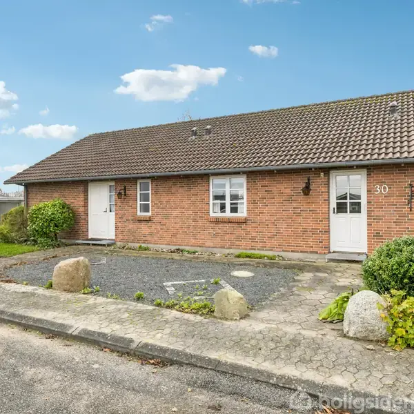 En murstensvilla med brunt tegltag står på en flisebelagt grund med omkransende buske. Dør markeret med nummeret 30. Skyfri himmel med hvide skyer i baggrunden.