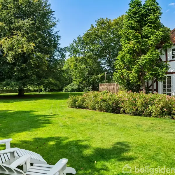 Hvide, træstole står på en velplejet græsplæne i en frodig have. Et bindingsværkshus med rødt tag ligger ved siden af, omgivet af høje træer og buske.