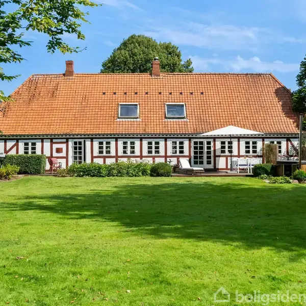 Et stort bindingsværkshus med rødt tegltag omgivet af velplejede græsplæner og træer. Huset har flere vinduer og en terrasse med havemøbler, der hviler under blå himmel.