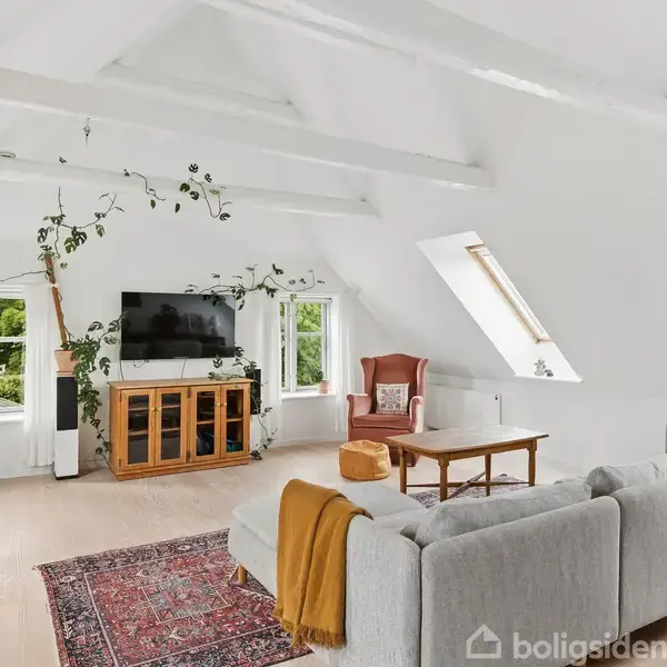 Stue med hvidmalede træbjælker, en grå sofa, fladskærms-TV på skænk, grønne planter og et rødt lænestol. Store vinduer og et skråt tag skaber lys og rummelighed.