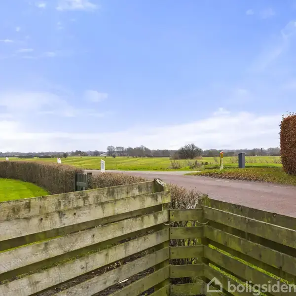 Træhegn står stille ved en grusvej, der fører ind i et åbent landsskab med grønne marker og blå himmel i baggrunden. En hæk løber langs vejen.