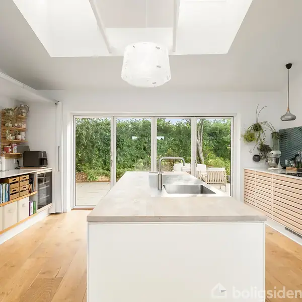 Et moderne køkken med en central ø, to håndvaske, og en bagvæg med blå fliser. Træskabe er langs væggen, og store glasdøre åbner til en terrasse med grøn udsigt.