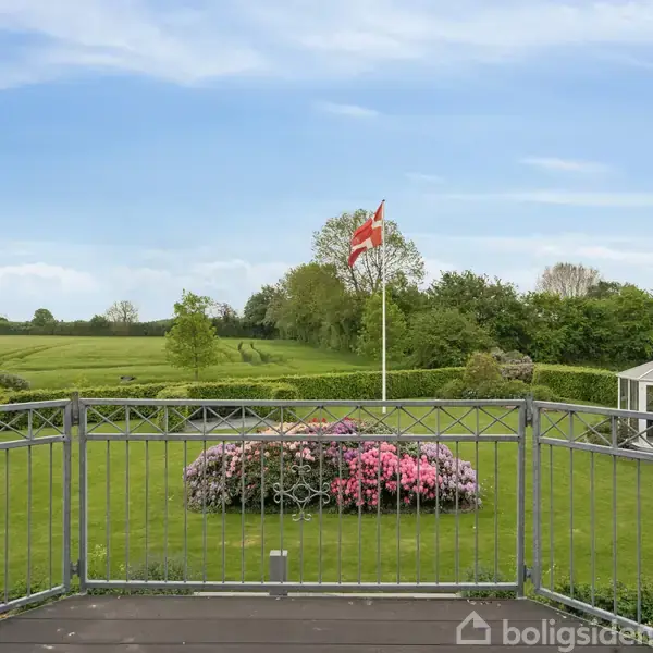 Et flag vajer i vinden over en grøn have med blomsterbed og en lysthus. Landskabet omkring er åbent, med marker og træer under en blå himmel.