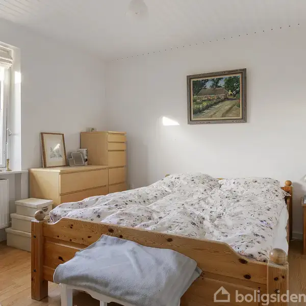 Sengen er centralt placeret og redt med blomstrede lagner. Ved siden af står trækommoder og natborde. Vinduet lader lys ind, og væggen har et naturmaleri.