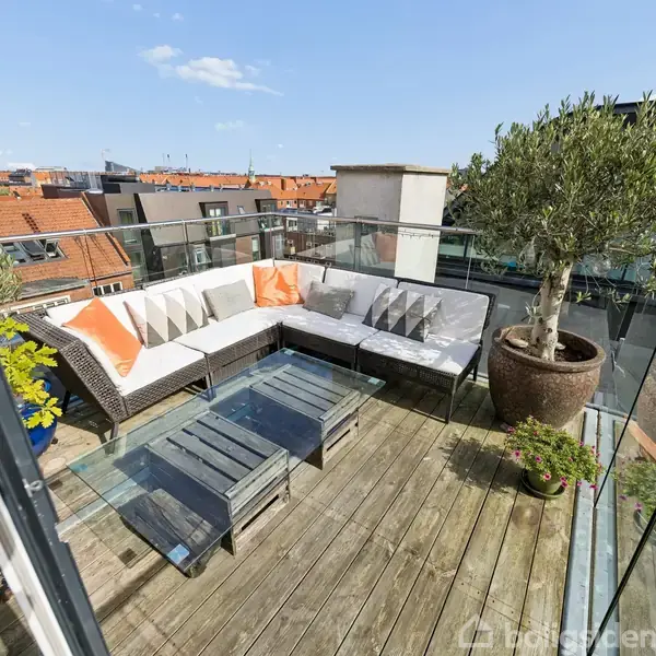 En L-formet sofa med farverige puder står på en træterrasse, flankeret af et glasbord og en stor plante. Terrassen er omgivet af glasrækværk med udsigt over byens tage.