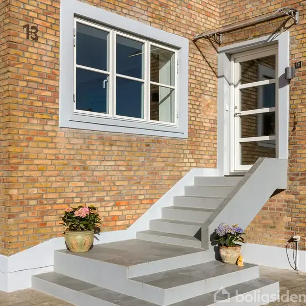 Trappe fører op til en hvid dør i en murstensfacade. En vinduesramme ses ved siden af trappen. To blomsterkrukker står ved trappens fod.