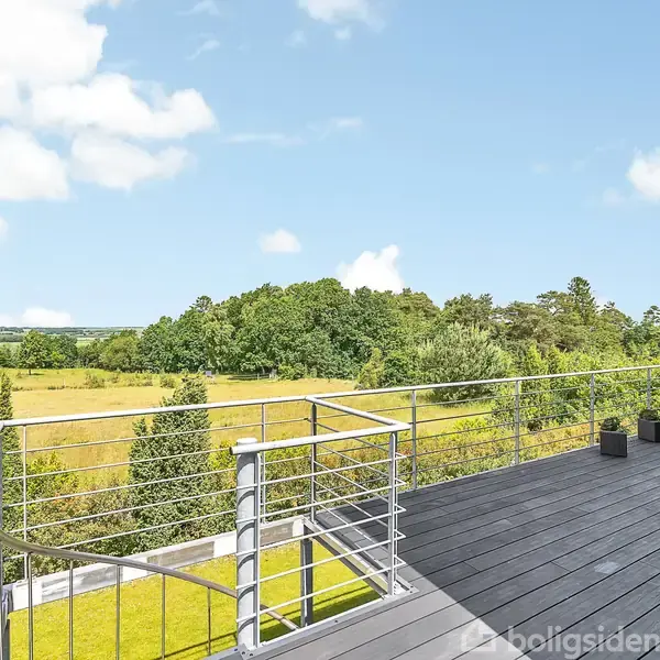 Stor terrasse med moderne rækværk strækker sig ud. Den har udsigt over en grøn landlig eng med træer i baggrunden og en blå himmel med få skyer. Tre plantekasser ses på terrassen.