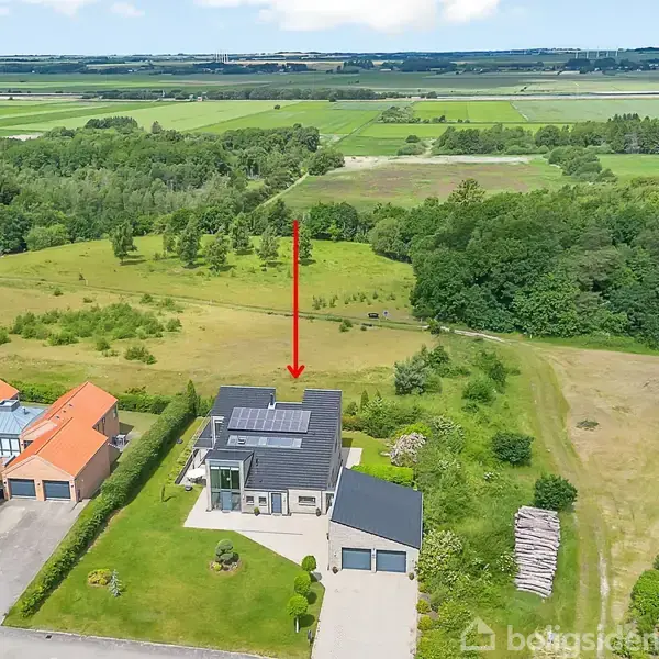Hus med grå tag og garage peges ud af en rød pil. Det er omgivet af grønne marker og skov i en landlig setting.