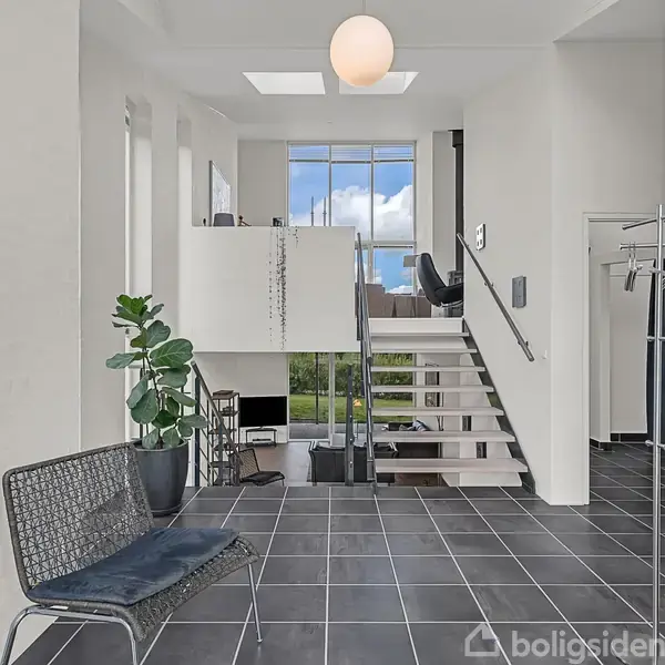 Stue med sorte klinkegulve; designstol og stueplante i forgrunden. I baggrunden fører en trappe til en øvre etage med store vinduer, der viser udsigt til blå himmel.