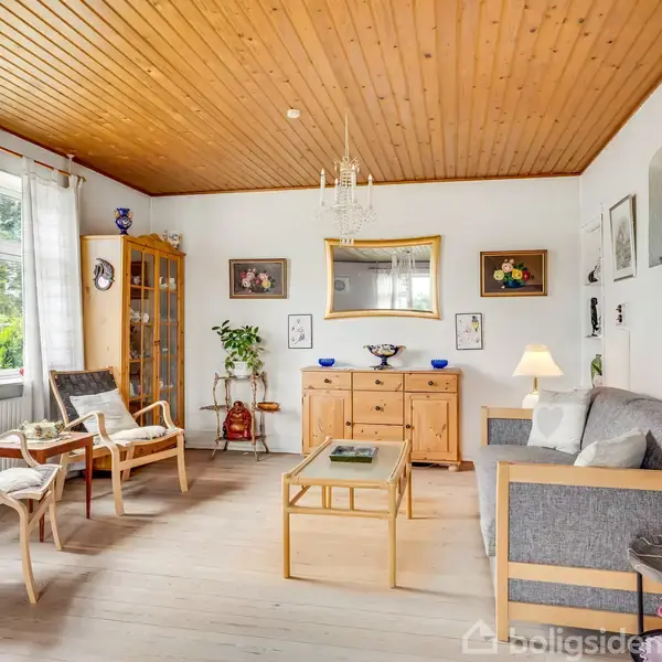 Stue med sofa og to stole står omkring et sofabord. Vitrineskab og kommode mod væggen. Trægulv og træloft, vindue med lyse gardiner. Lys og hyggelig atmosfære.