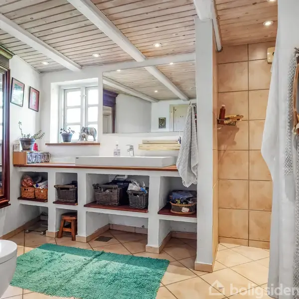 Et badeværelse med toilet. Håndklæder hænger ved en bruseniche. Grøn måtte ligger på et flisegulv, og træhylder under et spejl rummer kurve og beholder.