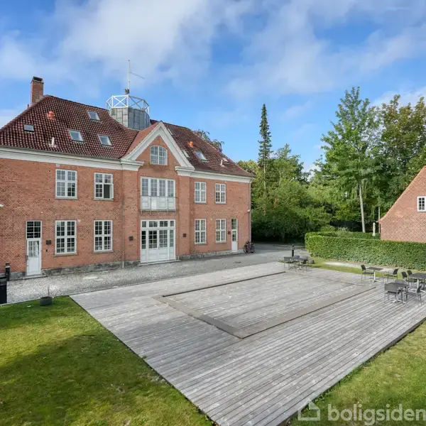 Stort rødt murstenshus i tre etager, med terrasseret trægård og havemøbler, omgivet af grønne træer. Himmel med spredte skyer i baggrunden.