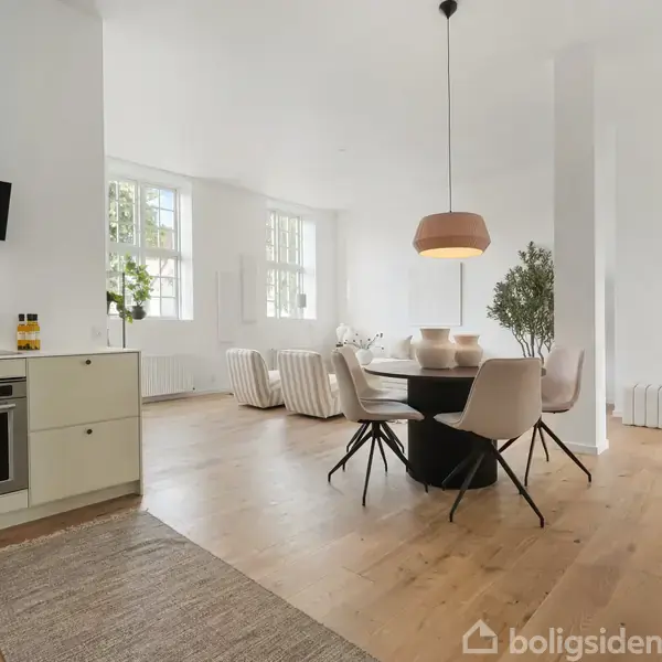 Et åbent køkken-alrum med moderne køkkenelementer til venstre. En sort rund spisebord med stole står i midten. Store vinduer i baggrunden lader lys ind i rummet.