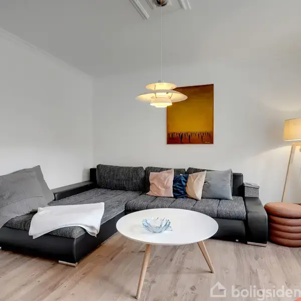 En grå sofa med farvede puder står i en stue. En hvid, rund sofabord står foran sofaen. En lampe og et kunstværk hænger på væggen.