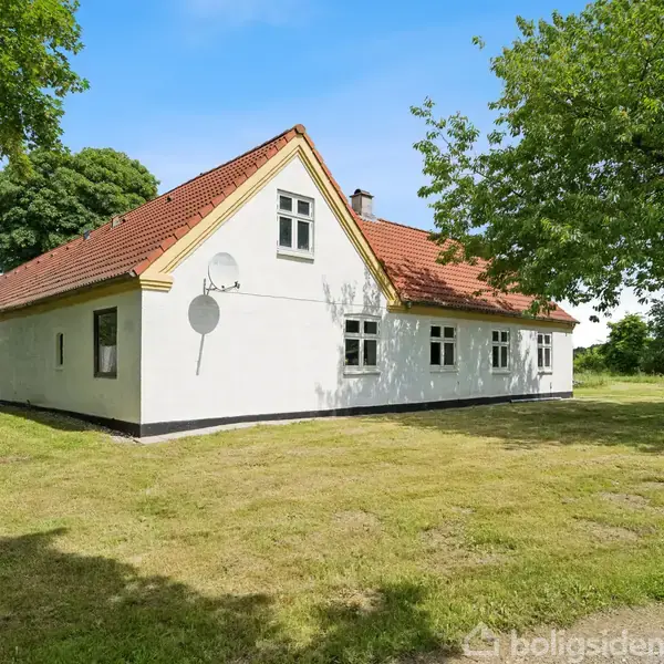 Et hvidt hus med rødt tegltag står omgivet af grønne træer og græsplæne. En parabolantennne er monteret på siden af huset. Klar blå himmel i baggrunden.