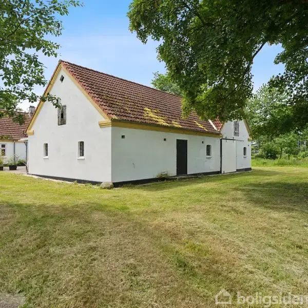 Hvidt hus med rødt tag står i en grøn have. Store træer omgiver huset, der har en rolig og landlig stemning. Græsplænen er velholdt, og himlen er klar.