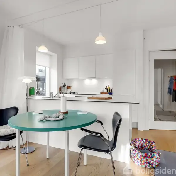 Et moderne køkken-alrum med hvide skabe og lys træbund, hvor et rundt, grønt bord med to sorte stole står i forgrunden. En farverig puf står ved siden af en åben dør.