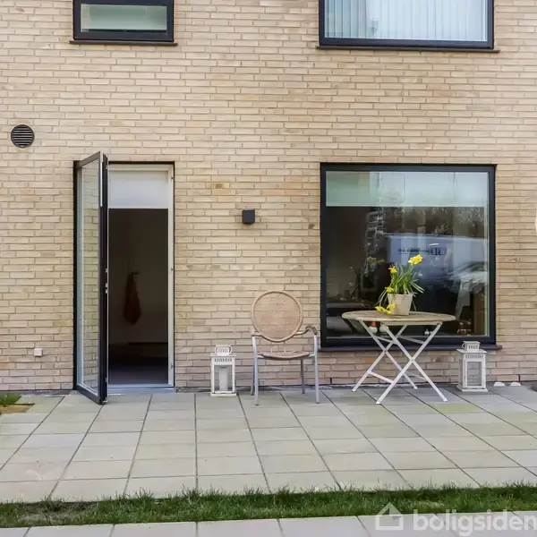 Dør står åben ved et moderne murstenshus. Et bord med blomster og en stol står på en flisebelagt terrasse. Paraplyparasol ses til højre, omkranset af græs og andre bygninger.