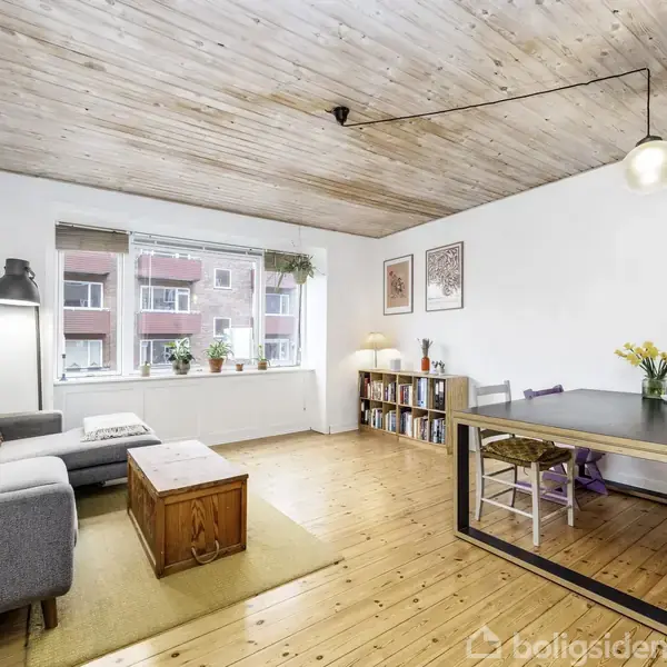 Stue med grå sofa og sofabord i træ står på et trægulv. Til højre er et spisebord med blomster. Rummet har hvidmalede vægge, træloft og stort vindue med udsigt til lejlighedsbygning.