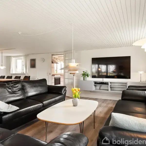 En stue med sorte lædersofaer placeret omkring et sofabord med blomster. I baggrunden ses et stort tv på en hvid reol og et spisebord i en åben, lys indretning.