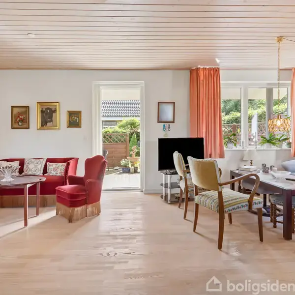 Stue med møbler i klassisk stil, herunder sofaer, borde og stole. Vinduer med røde gardiner lukker lys ind fra haven udenfor. Væggene er dekoreret med malerier.