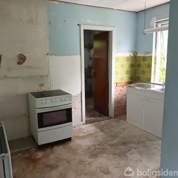 En ovn står ubenyttet i et slidt køkken med afskallede vægge. Et hvidt skab og en vask findes ved siden af et vindue.