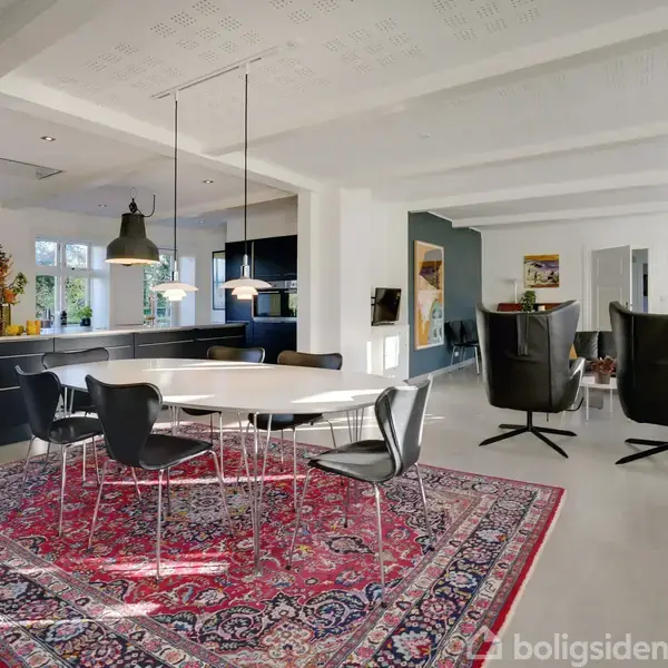Langbord med sorte stole er placeret på et rødt orientalsk tæppe i en modernistisk stue, oplyst af loftslamper. Køkkenet med hvide skabe ses i baggrunden ved siden af store vinduer.