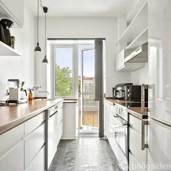 Et lyst køkken med hvide skabe og træbordplader. To lamper hænger fra loftet. I baggrunden ses en åben dør til en balkon med udsigt til træer og bygninger.