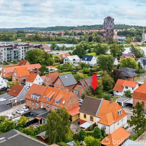Et boligområde med røde og orange tage. En rød pil peger mod et specifikt hus blandt tætstående huse. I baggrunden ses en sø og bygninger.