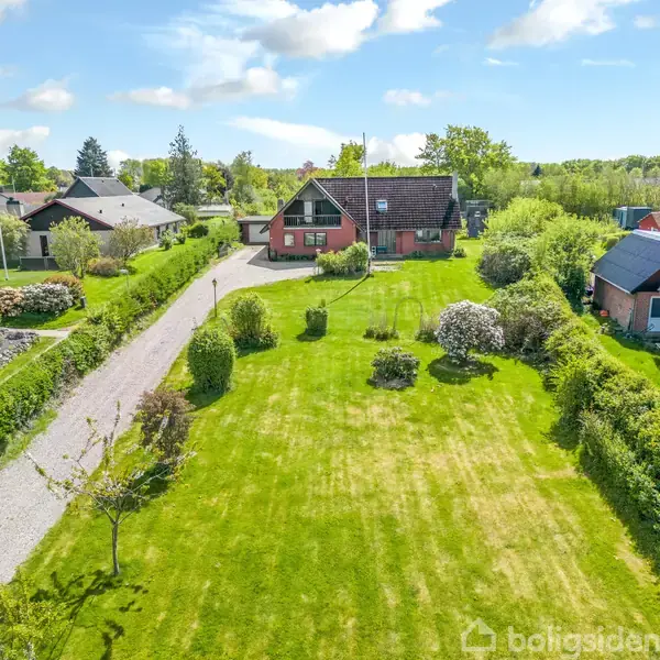 Rødt hus med skråt tag står for enden af en lang, velplejet græsplæne omgivet af buske og træer. Grusvej leder op til huset, der ligger i et frodigt, landligt område.