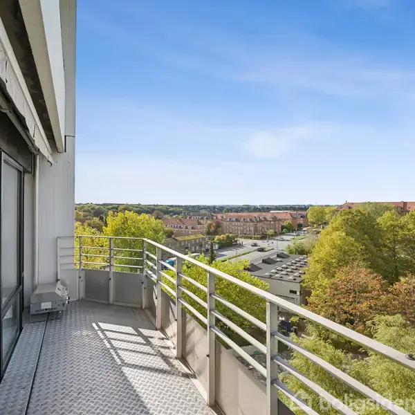 Balkon med metalrækværk, vender ud mod en udsigt over by og træer, under en klar blå himmel.