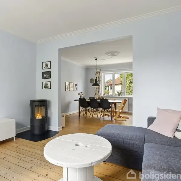 En stue med en grå sofa og et rundt bord udført i lyst træ står tæt ved en væg. Til højre ses en åbning til en spisestue med mørke stole omkring et bord. Et fjernsyn er placeret på et hvidt skab, og en brændeovn er tændt på gulvet. Væggene er lyseblå, og gulvet er træ.