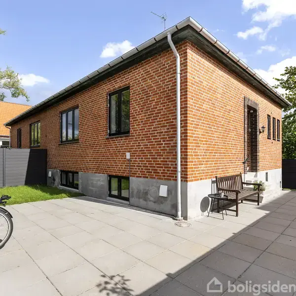 Rødt murstenshus står stille i solfyldt have med flisebelagt terrasse. En cykel er parkeret ved siden af en bænk under et stort vindue.