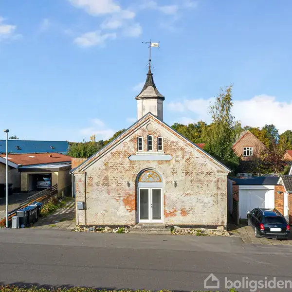 En ældre bygning med slidt murstensfacade og et lille tårn med en vindfløj. Bygningen står på en stille vej mellem to moderne huse med træer i baggrunden.