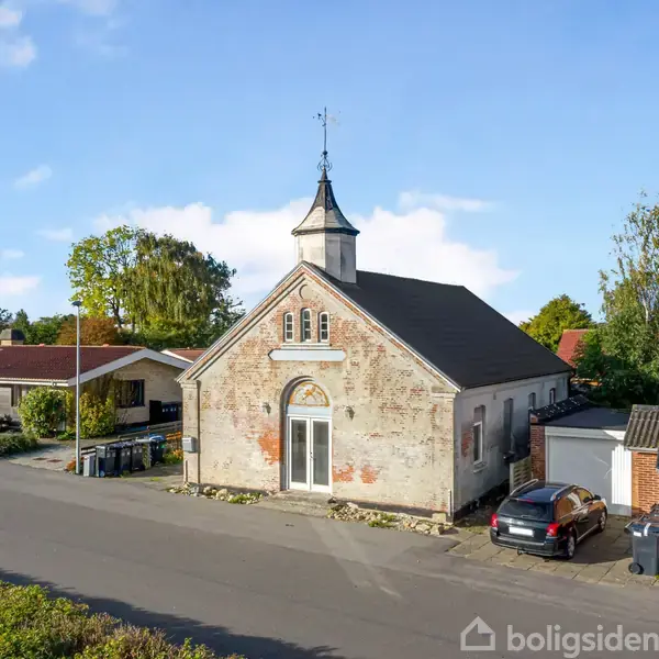 En ældre bygning med tårn står midt på en villavej højt beliggende omgivet af huse og træer under en blå himmel.
