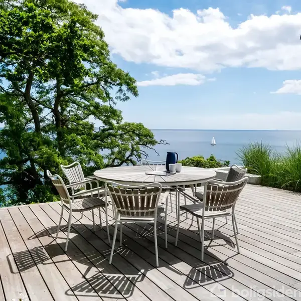 Rundt havebord med fire stole står på en træterrasse. I baggrunden ses havudsigt med en sejlbåd, omkranset af grønne træer og blå himmel med skyer.