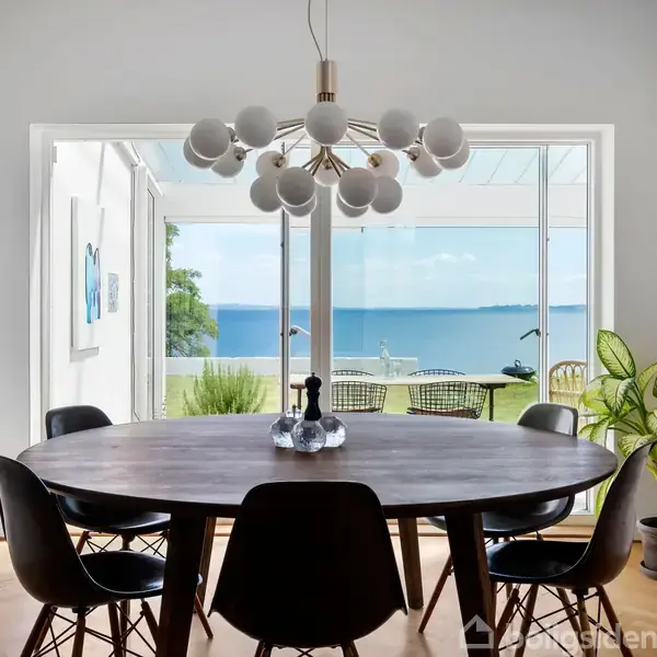 Rundt bord omgivet af sorte stole står i et lyst rum med udsigt over en terrasse og havet. En moderne lysekrone hænger over bordet, og planter ses i rummet.