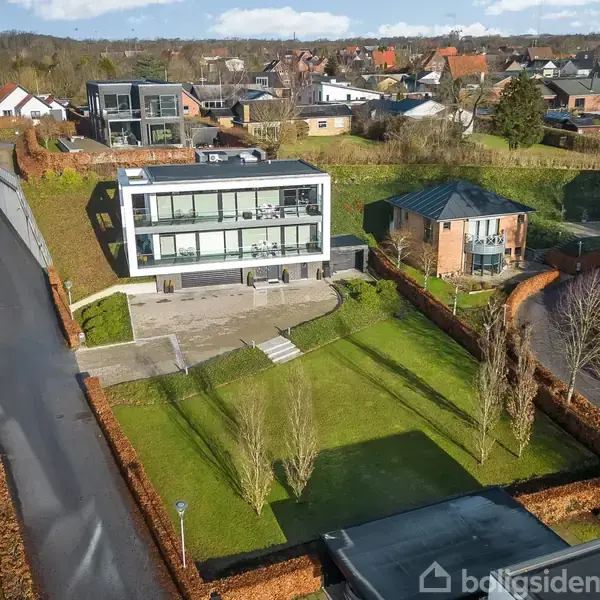 Moderne, hvidt hus med store vinduer står på en velplejet græsplæne. Træer omgiver huset, og en brolagt indkørsel leder op til det. Omgivet af flere moderne bygninger i et boligområde.