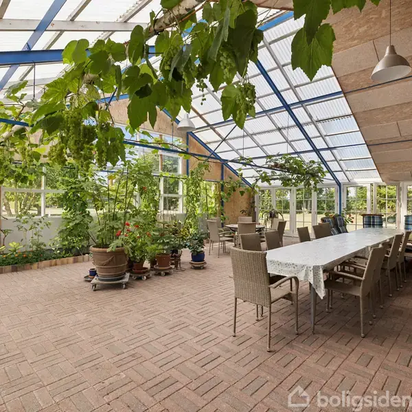 Langt bord med stole under gennemsigtigt tag, omgivet af grønne planter i pottedyrkning; luftigt rum med udsigt til grønt udenfor.