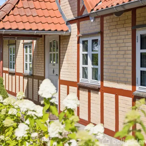 Hus med gule mursten og rødt tag står fredfyldt i solen; omkranset af blomstrende buske i en velplejet have.