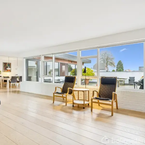 To lænestole står side om side med et lille rundt bord imellem sig ved store vinduer, der giver udsigt til en terrasse. I baggrunden ses et spisebord ved en sort væg.