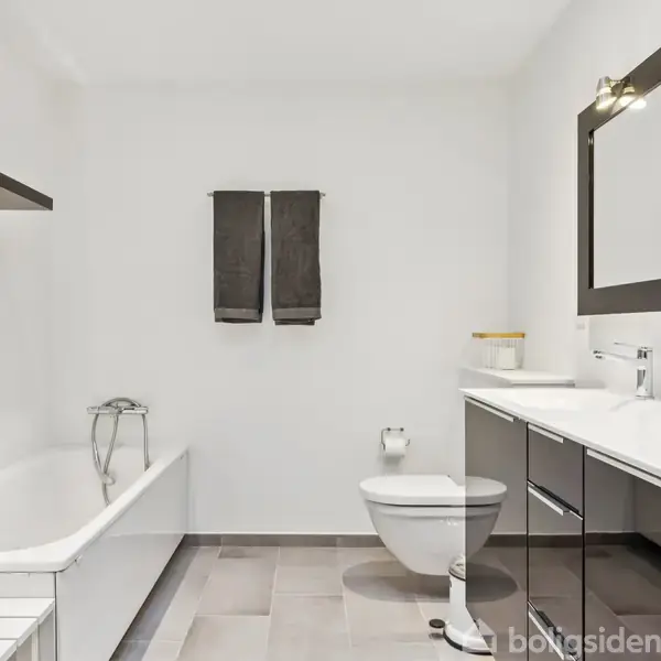 Badeværelse med hvidt badekar til venstre og moderne vaskeskab til højre, med spejl og håndvask. To grå håndklæder hænger på væggen. Enkel og moderne indretning.