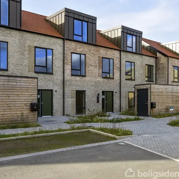 Rækkehuse med moderne, fladskårne facader, står side om side, hver med store vinduer og grønne døre. Fortovsfliser og små græsarealer ligger foran bygningerne.